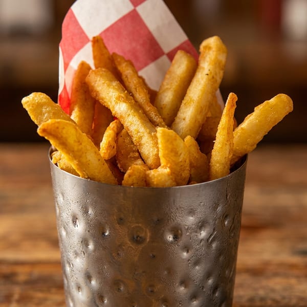 Golden french fries in a hammered metal cup with red and white checkered paper on a wooden table.