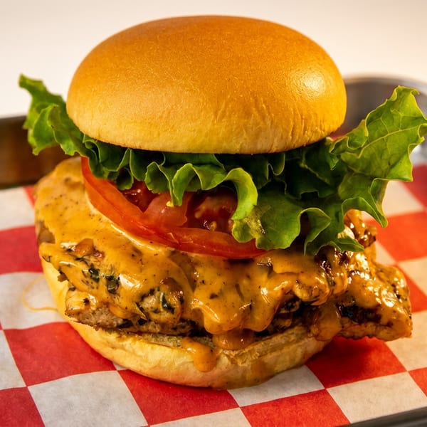 Chicken sandwich with lettuce, tomato, and creamy sauce on a bun, served on checkered paper.
