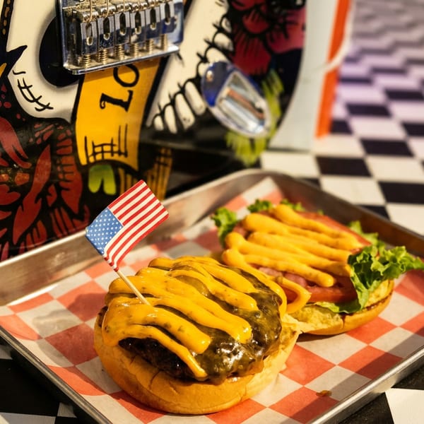 Two cheeseburgers on a tray with an American flag, beside a decorated guitar and checkered table.