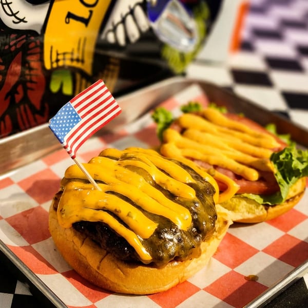 Two cheeseburgers with yellow cheese strips and an American flag on a tray, on a checkered table.